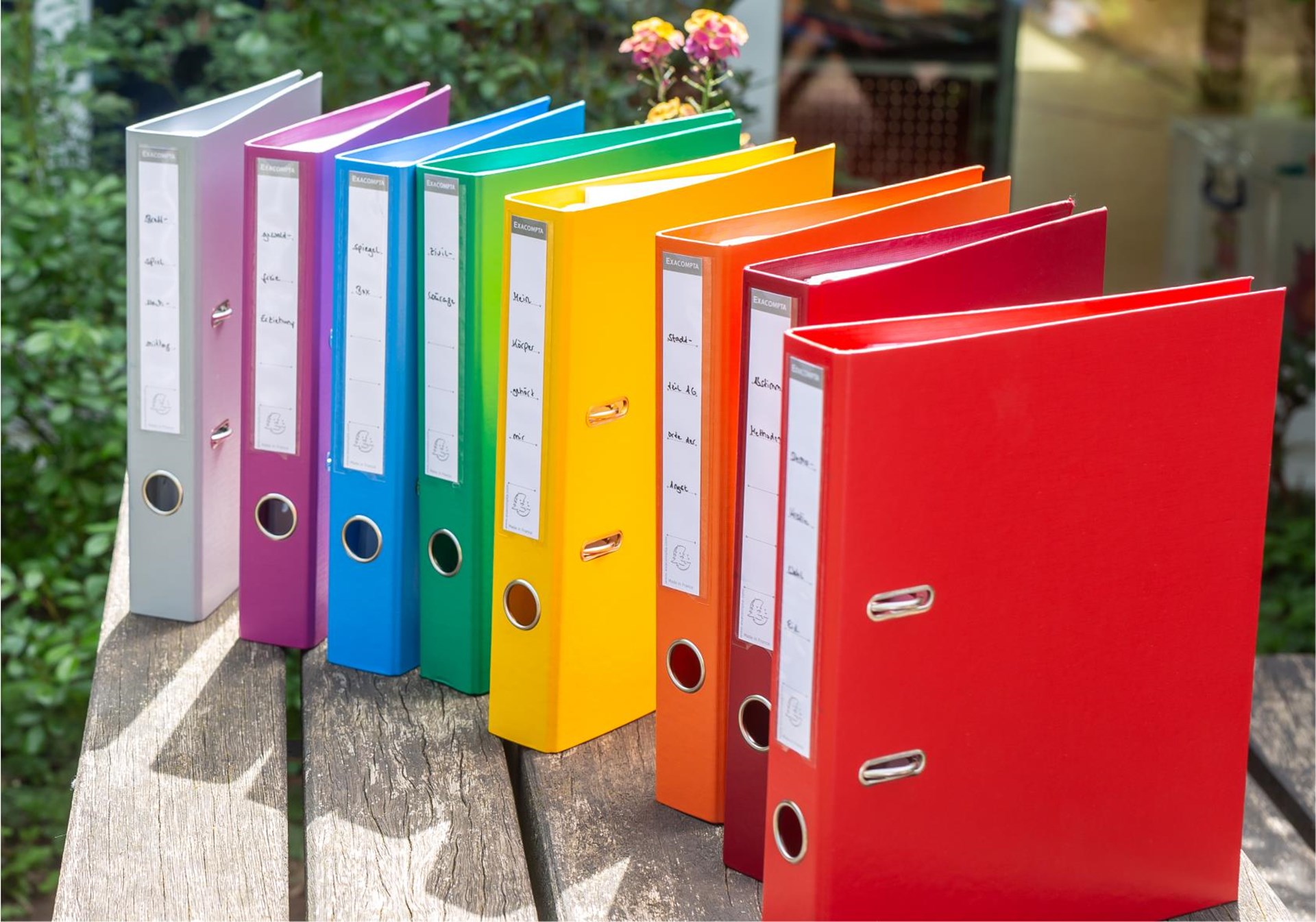 Eine Reihe von farbenfrohen Ordnern, angeordnet wie ein Regenbogen, stehen auf einem Holztisch vor einem unscharfen Hintergrund.